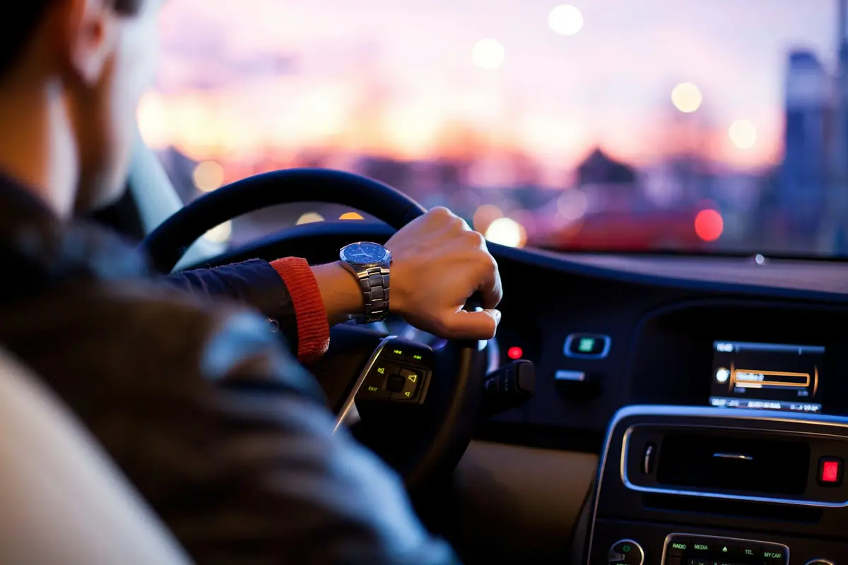 Driver's hands on the wheel at dusk, city lights blurred through the windshield
